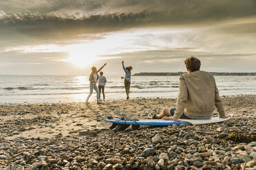 Young man on surfboard watching friends making soap bubbles on the beach at sunset - UUF11667