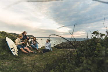 Happy friends with surfboard and drinks socializing at the coast at sunset - UUF11673