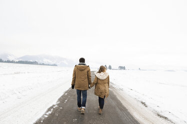 Rückenansicht eines jungen verliebten Paares, das Hand in Hand auf einer Landstraße in einer verschneiten Landschaft spazieren geht - HAPF02056