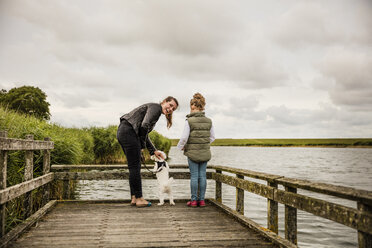 Mutter und Tochter stehen auf einem Steg an einem See mit Hund - MOEF00108