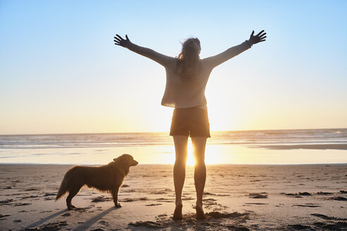 Portugal, Algarve, Frau mit Hund hebt Arme am Strand bei Sonnenuntergang - JRF00341