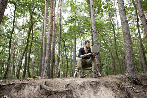 Mann sitzt auf einem selbstgebauten Holzstuhl im Wald und benutzt einen Laptop - MFRF01048