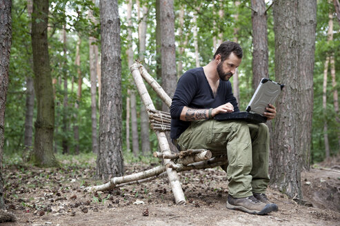Mann sitzt auf einem selbstgebauten Holzstuhl im Wald und benutzt einen Laptop - MFRF01050