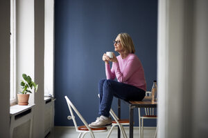 Mature woman at home with cup of coffee looking out of window - RBF05871