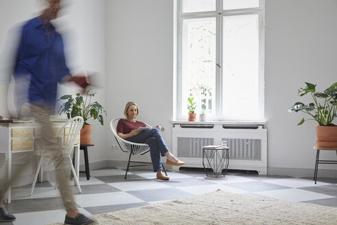 Mature woman reading magazine at home with man passing by - RBF05894