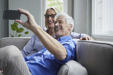 Glückliches reifes Paar sitzt auf der Couch zu Hause und macht ein Selfie - RBF05900