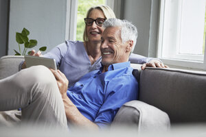 Happy mature couple sitting on couch at home sharing tablet - RBF05901