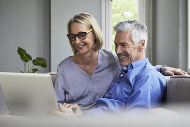 Happy mature couple sitting on couch at home sharing laptop - RBF05902