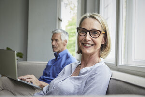 Portrait of smiling mature woman on couch with man in background using tablet - RBF05904
