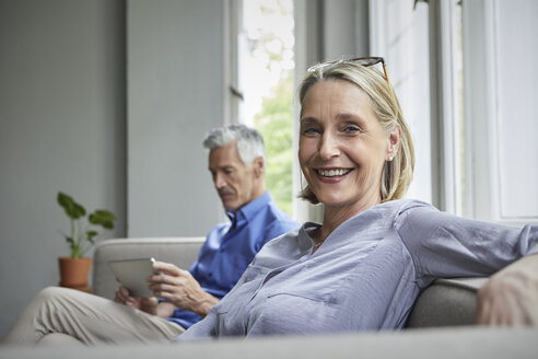 Porträt einer lächelnden reifen Frau auf einer Couch mit einem Mann im Hintergrund, der ein Tablet benutzt - RBF05905