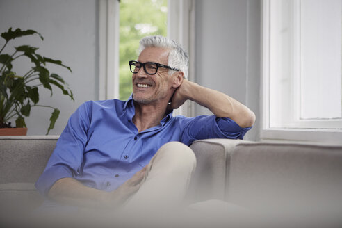 Porträt eines lachenden reifen Mannes, der zu Hause auf der Couch sitzt - RBF05945
