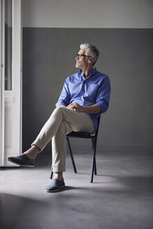 Pensive mature man sitting on chair at home looking at distance - RBF05953