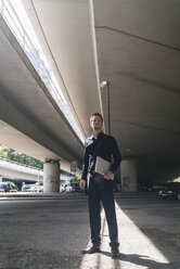 Businessman standing at underpass holding laptop - KNSF02473
