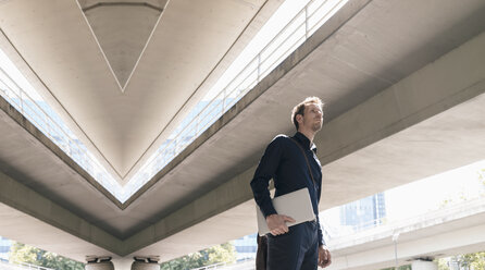 Businessman standing at underpass holding laptop, composite - KNSF02475