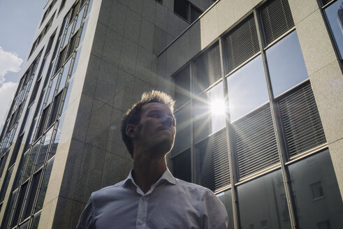 Businessman standing outdoors with sun reflecting in office building - KNSF02479