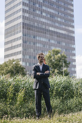 Lächelnder Geschäftsmann steht auf einem Feld vor einem Bürogebäude - KNSF02519