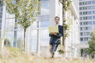 Geschäftsmann lehnt an einem Baum vor einem Bürogebäude und benutzt einen Laptop - KNSF02524