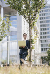 Geschäftsmann lehnt an einem Baum vor einem Bürogebäude und benutzt einen Laptop - KNSF02525