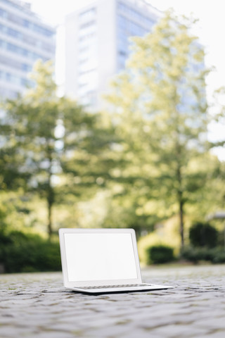 Laptop auf Kopfsteinpflaster im Park mit Bürogebäuden im Hintergrund, lizenzfreies Stockfoto