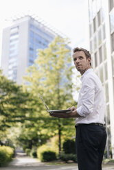 Businessman using laptop in front of office building - KNSF02535