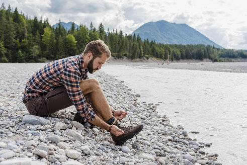 Germany, Bavaria, hiker sitting at riverside tying his shoe - DIGF02807