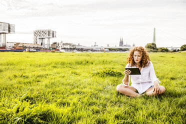 Germany, Cologne, young woman sitting on meadow looking at tablet - FMKF04384