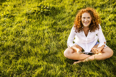 Porträt einer glücklichen jungen Frau mit Tablet, die sich auf einer Wiese entspannt - FMKF04386