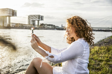 Germany, Cologne, young woman sitting at riverside in the evening taking selfie with cell phone - FMKF04393