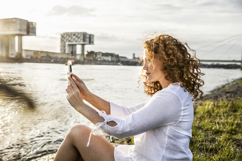 Deutschland, Köln, junge Frau sitzt abends am Flussufer und macht ein Selfie mit Handy - FMKF04393
