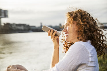 Germany, Cologne, portrait of laughing young woman on the phone sitting at riverside in the evening - FMKF04395