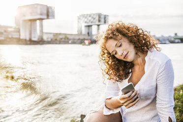 Deutschland, Köln, Porträt einer jungen Frau, die am Flussufer sitzt und auf ihr Mobiltelefon schaut - FMKF04397