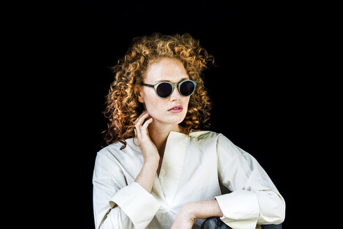 Portrait of stylish young woman with curly red hair wearing sunglasses in front of black background - FMKF04403