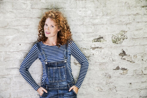 Portrait of smiling young woman wearing denim dungarees leaning against white brick wall - FMKF04412