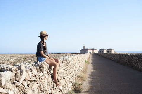 Spanien, Menorca, Einzelreisender auf Natursteinmauer sitzend mit Blick auf die Aussicht, lizenzfreies Stockfoto
