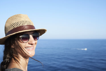 Portrait of smiling woman wearing straw hat and sunglasses in front of the sea - IGGF00155