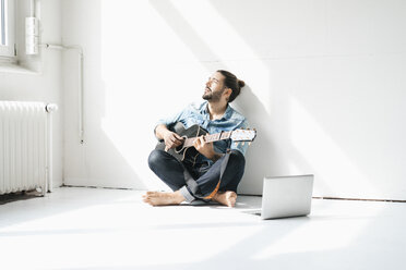 Mann sitzt mit Laptop auf dem Boden in einem Loft und spielt Gitarre - JOSF01527