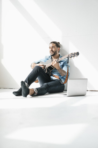 Singender Mann sitzt mit Laptop auf dem Boden und spielt Gitarre, lizenzfreies Stockfoto