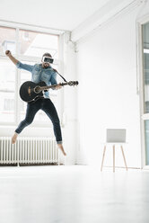 Mann mit Gitarre und Virtual-Reality-Brille springt in einem Loft in die Luft - JOSF01533