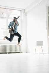 Mann mit Gitarre und Virtual-Reality-Brille springt in einem Loft in die Luft - JOSF01534