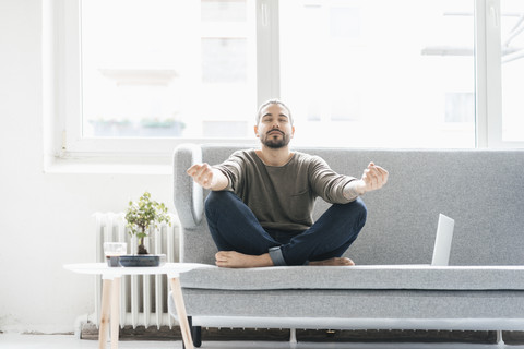 Porträt eines Mannes mit geschlossenen Augen, der auf der Couch sitzt und Yogaübungen macht, lizenzfreies Stockfoto