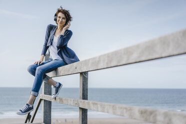 Frau sitzt auf einem Geländer am Strand und hört Musik - KNSF02544