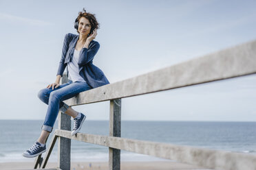 Woman sitting on railing at the beach listening to music - KNSF02544