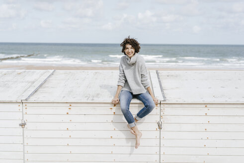Lächelnde Frau sitzt auf einer Holzkiste am Strand - KNSF02583