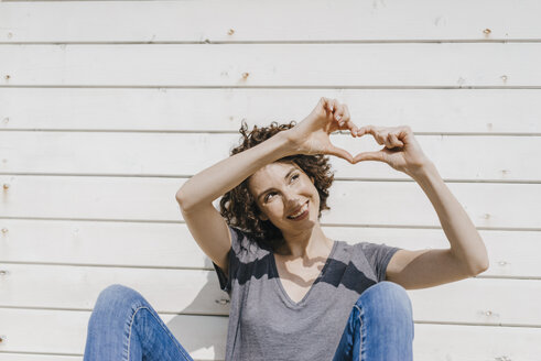 Woman leaning against wooden wall shaping heart with her hands - KNSF02593
