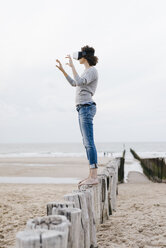 Woman standing on wooden stake on the beach wearing VR glasses - KNSF02622