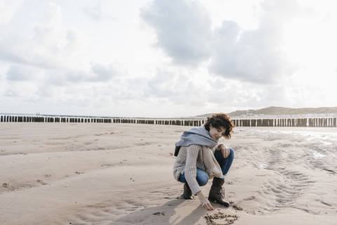 Frau hockt am Strand und zeichnet ein Herz in den Sand, lizenzfreies Stockfoto