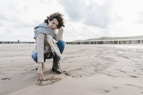 Woman crouching on the beach drawing a heart in the sand stock photo