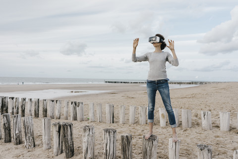 Frau steht auf Holzpfählen am Strand und trägt eine VR-Brille, lizenzfreies Stockfoto