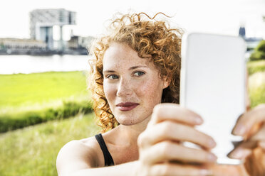 Germany, Cologne, portrait of freckled young woman taking selfie with cell phone - FMKF04432