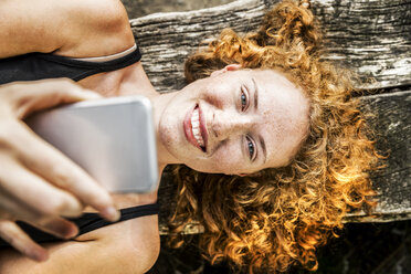 Porträt einer rothaarigen jungen Frau, die auf einer Bank liegt und ein Mobiltelefon benutzt - FMKF04436